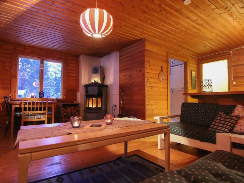 Cozy wooden interior with sofas, table, dining area, and fireplace, lit by warm lighting.