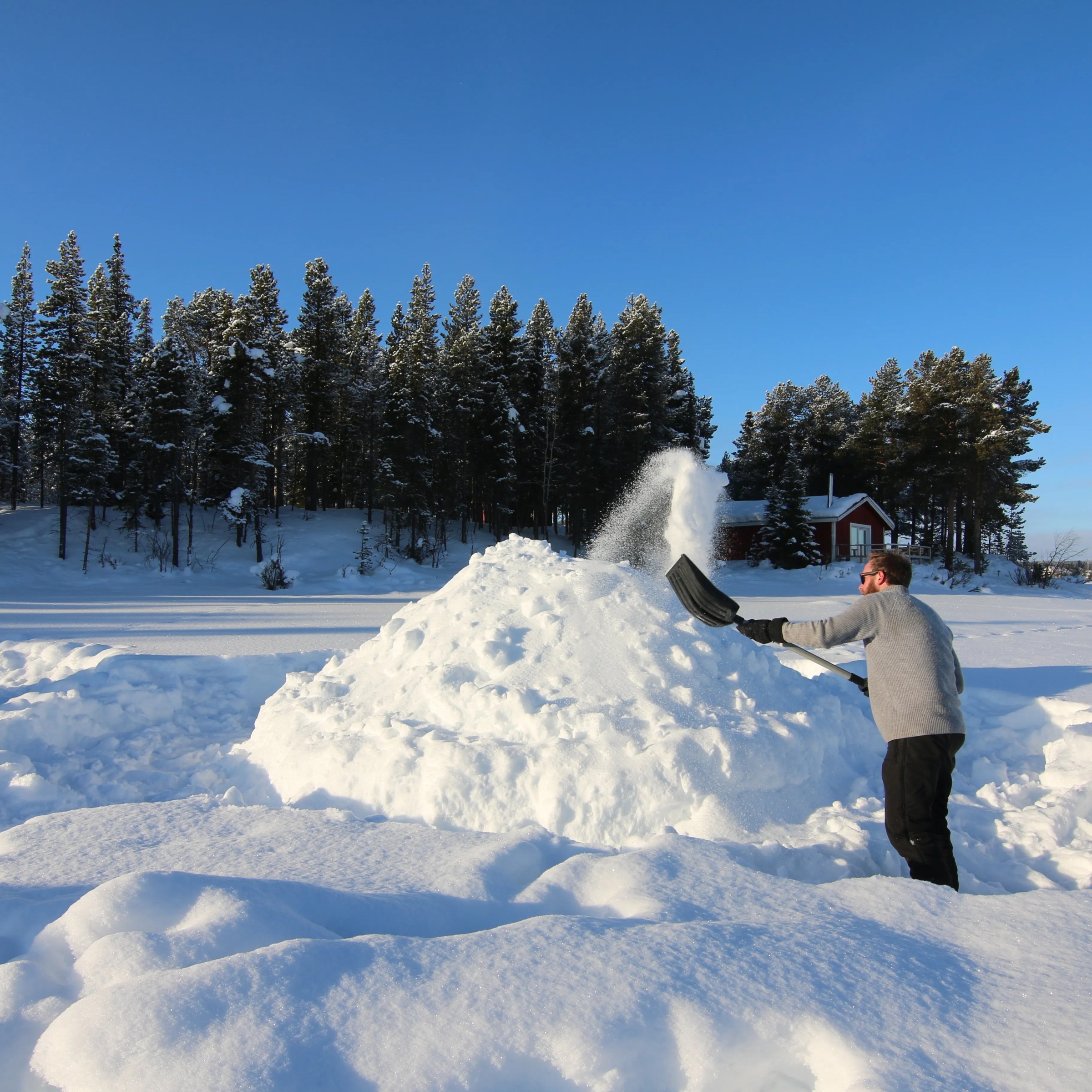 Person shoveling snow into a pile outdoors with snowy trees and a cabin in the background.