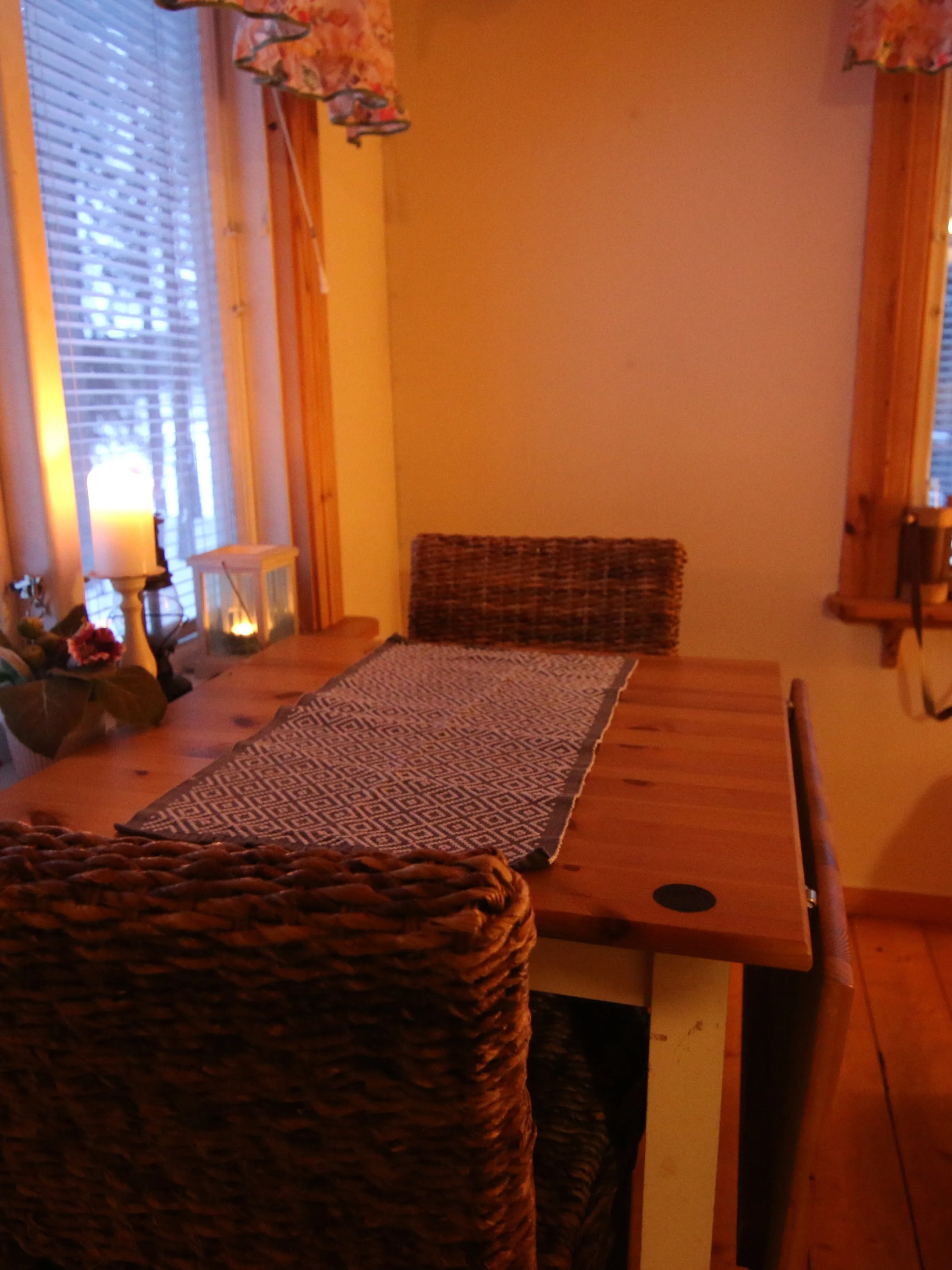 Cozy room with wooden table, woven chairs, and candles by a window with snowy view.
