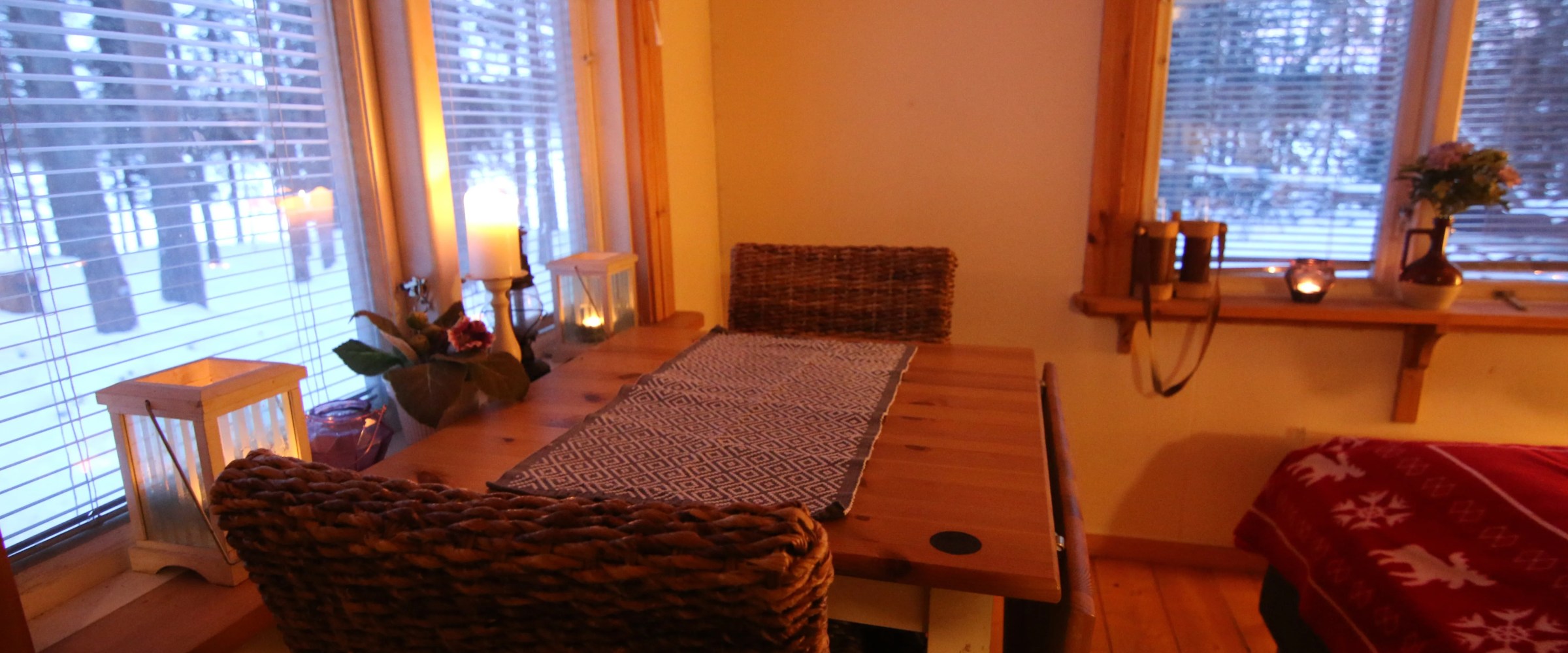 Cozy room with wooden table, woven chairs, and candles by a window with snowy view.