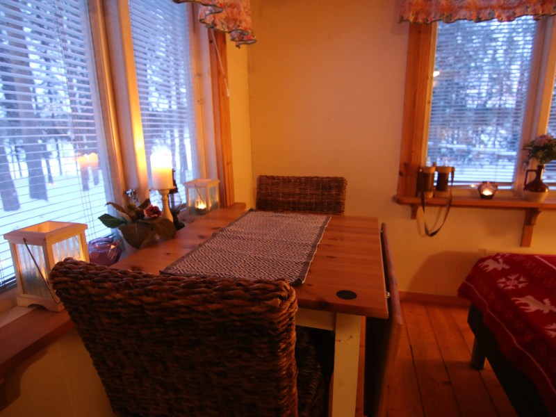 Cozy room with wooden table, woven chairs, and candles by a window with snowy view.