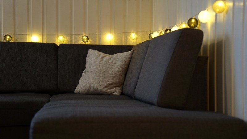 Gray corner sofa with a beige pillow, decorated with string lights on a paneled wall.