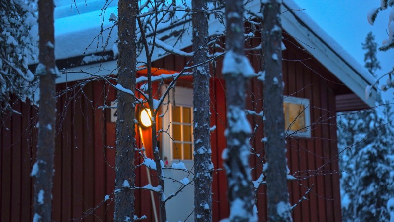 Snow-covered trees in front of a warmly lit red cabin at dusk.