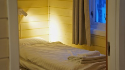 Cozy wooden room with a single bed, soft lighting, and folded towels by a curtained window.