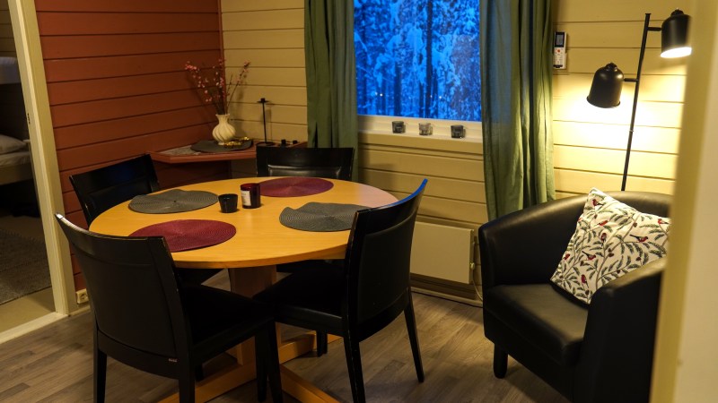 Cozy dining area with round table, four chairs, and lamp-lit armchair near window at dusk.