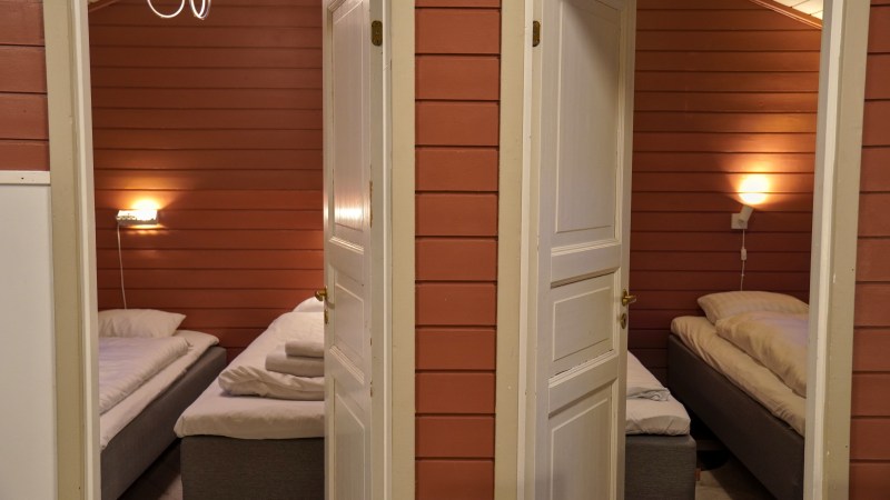 Two open doors reveal beds in adjacent cozy wooden rooms with wall lights.