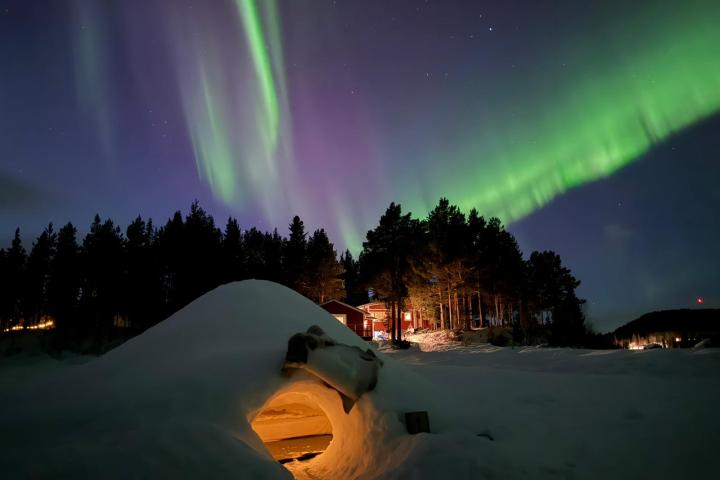 Aurora Borealis over snowy landscape with igloo and red cabin at night.
