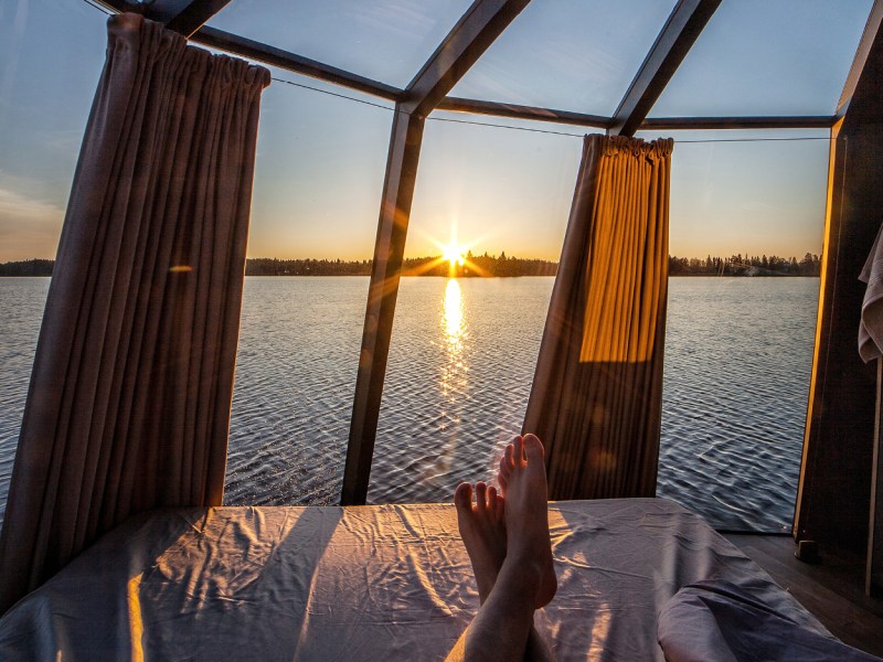 View of lake sunset from bed in glass-walled room with curtains.