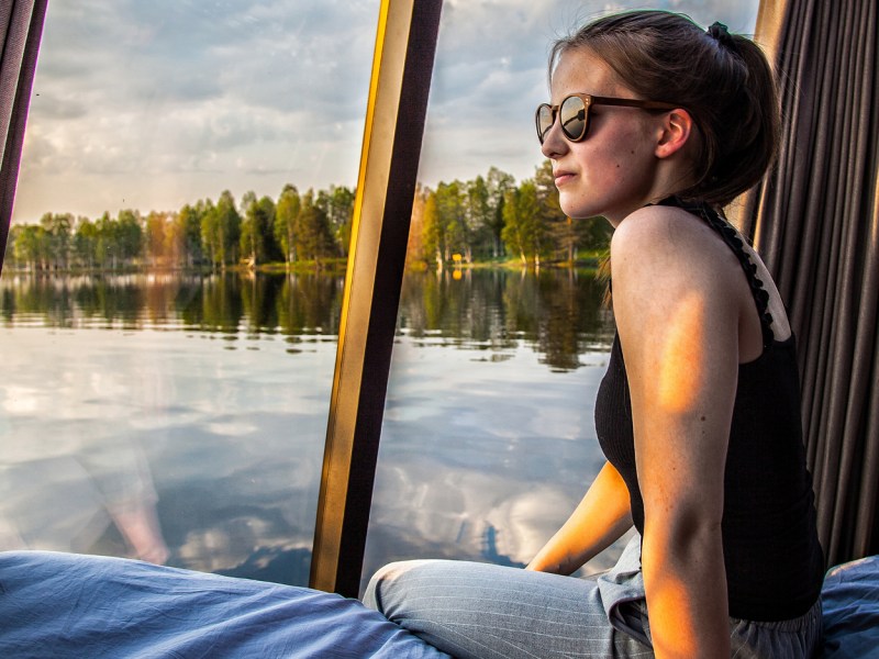 Woman in sunglasses sitting by a large window with lake and trees outside.