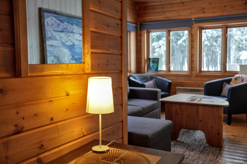 Cozy wooden cabin interior with lamp, mirror, sofa, and snow-covered trees visible outside windows.