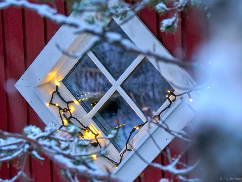 Diamond window with snow-covered frame and holiday lights on a red building.