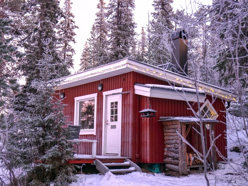 Red cabin in snowy forest with string lights, logs, and chimney.