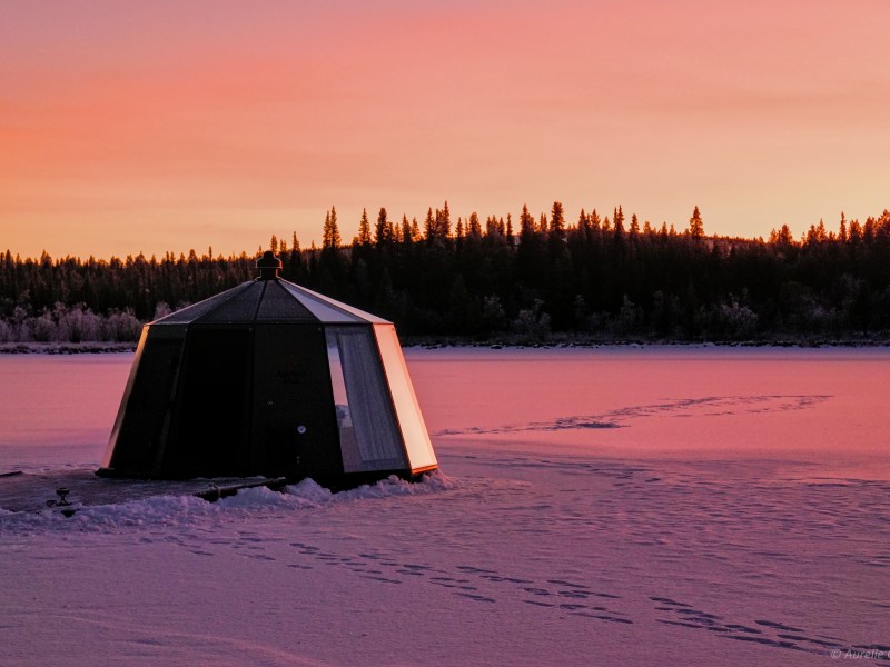 Snowy landscape with a small, dark structure and forest under a pink sunset sky.