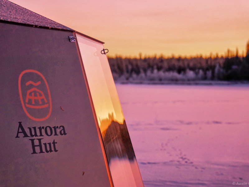 Aurora hut at sunset with snowy landscape and forest in background.