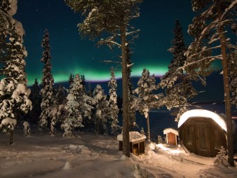 Snowy forest with Northern Lights and a small cabin under starry night sky.