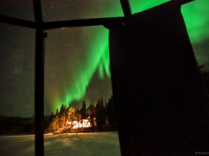 Green aurora borealis over snowy landscape and lit trees viewed through a window.
