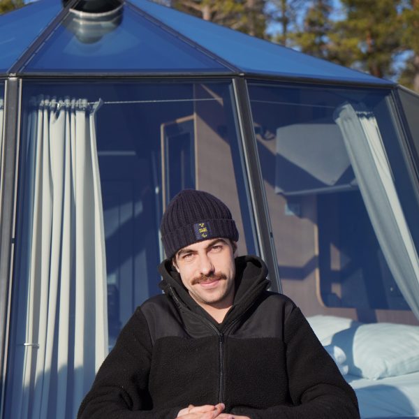 Person in winter clothes sitting outside a glass cabin, surrounded by trees.