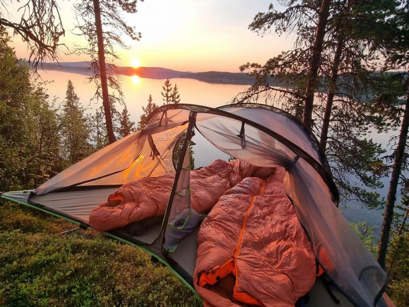 Tent suspended among trees overlooking a lake at sunset with orange sleeping bags inside.