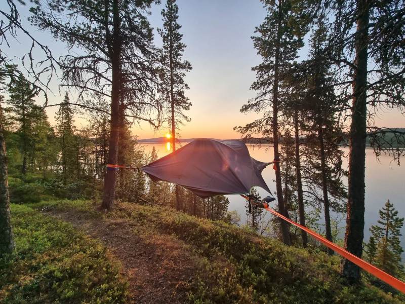 Tent suspended between trees by a lake at sunset.