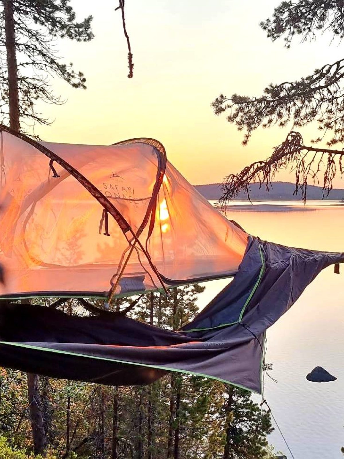 Tent suspended between trees overlooking a lake and sunset.