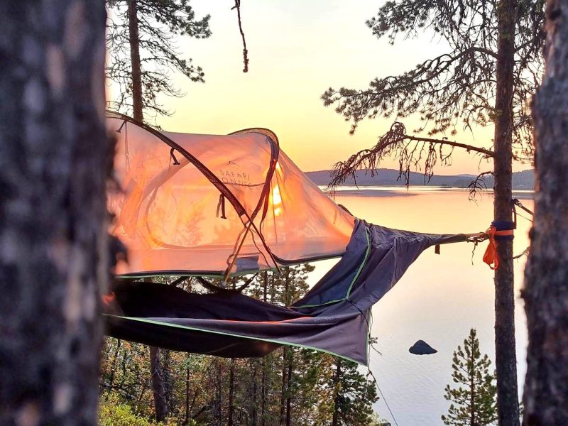 Tent suspended between trees overlooking a lake and sunset.
