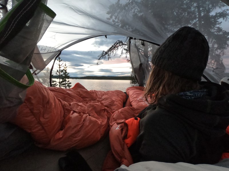Person in tent looking at lake and trees during sunset.