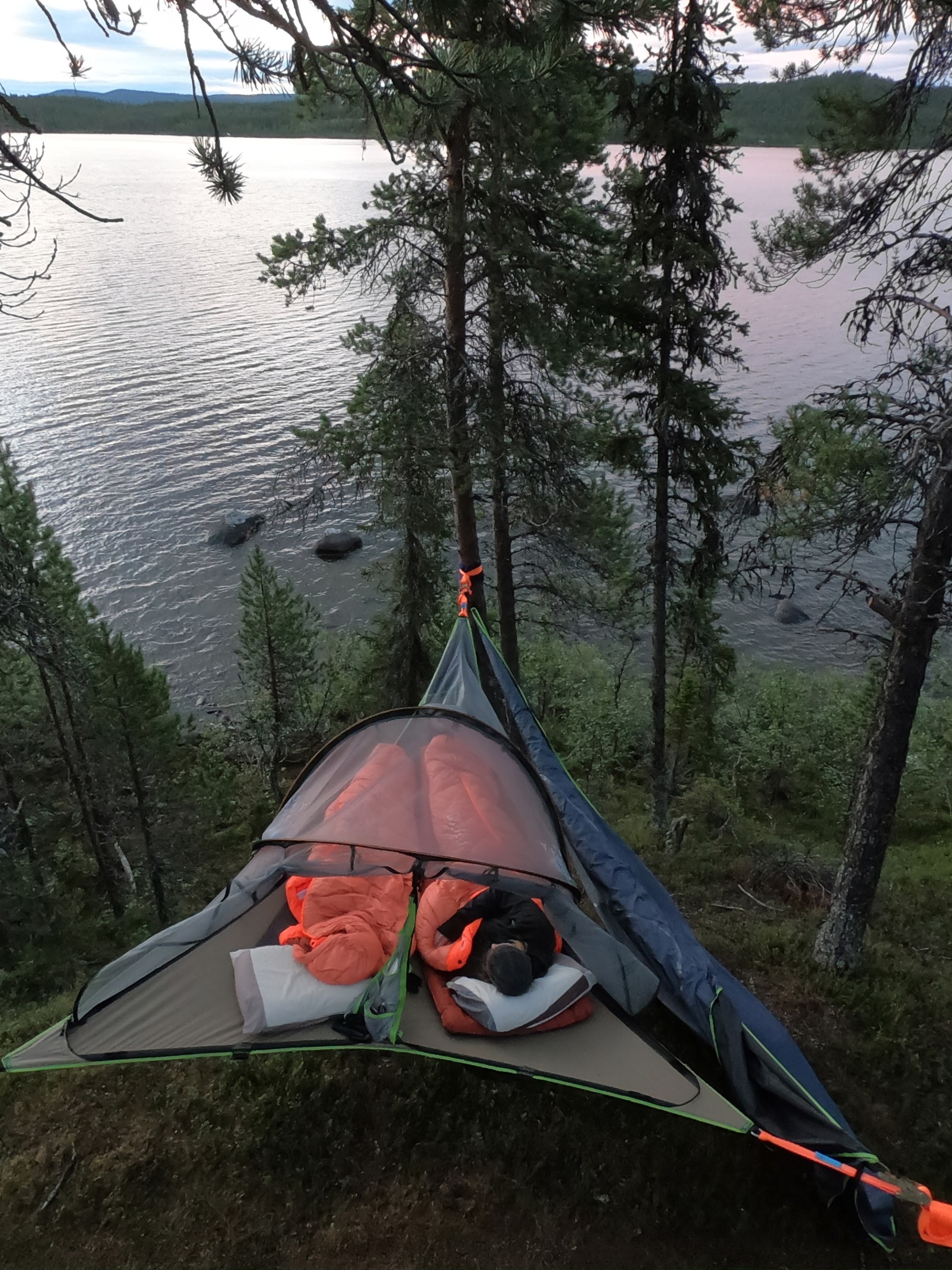 Suspended tent among trees with view of a lake at dusk.