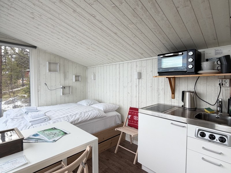 Small cabin with bed, table, chair, and kitchenette under slanted ceiling.