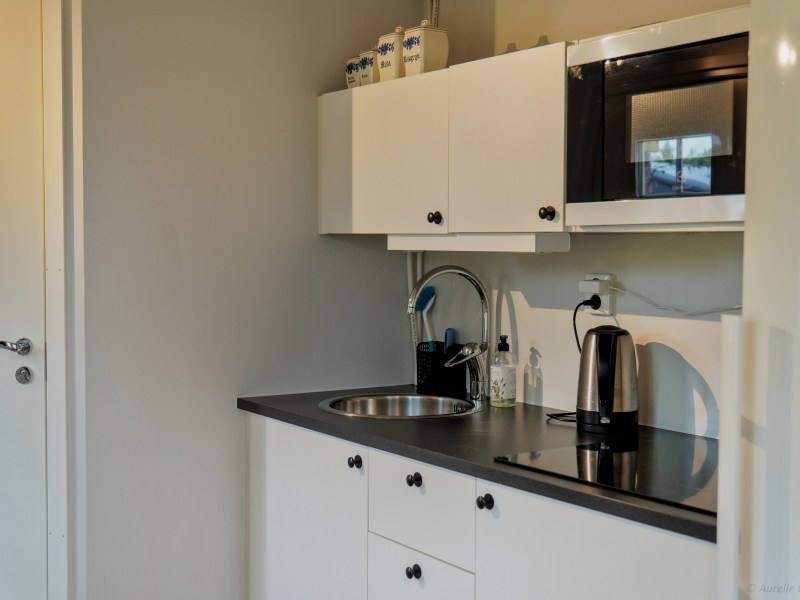 Compact kitchen with white cabinets, a microwave, sink, and kettle on a black countertop.