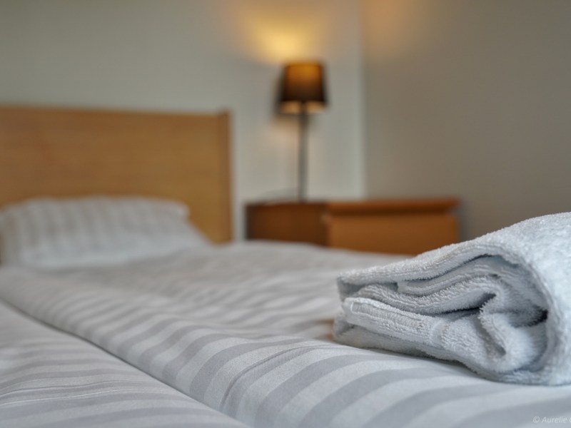 Neatly made bed with striped sheets and folded towel, lamp in background.
