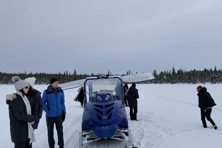 People standing near a blue snowmobile on a snowy landscape with trees in the background.